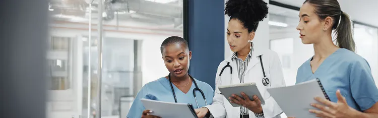 A group of medical professionals in white coats and scrubs engage in a discussion in an office setting.