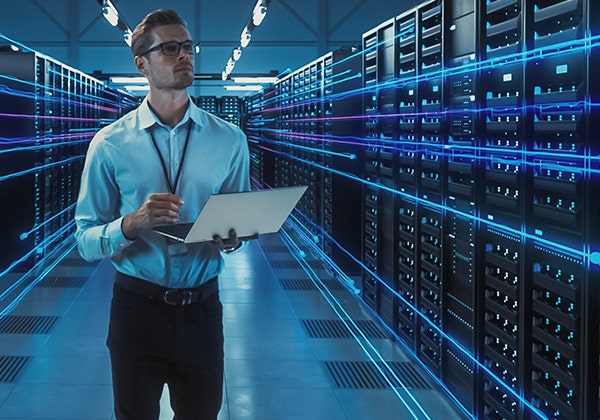 A male employee holding his laptop looking a row of datacenter.