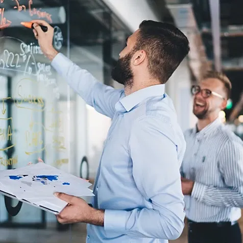 Man holding some papers in one hand and writing on a board with a marker with the other while another man watches, smiling