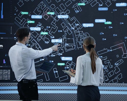Two individuals stand before a large screen displaying a detailed map.