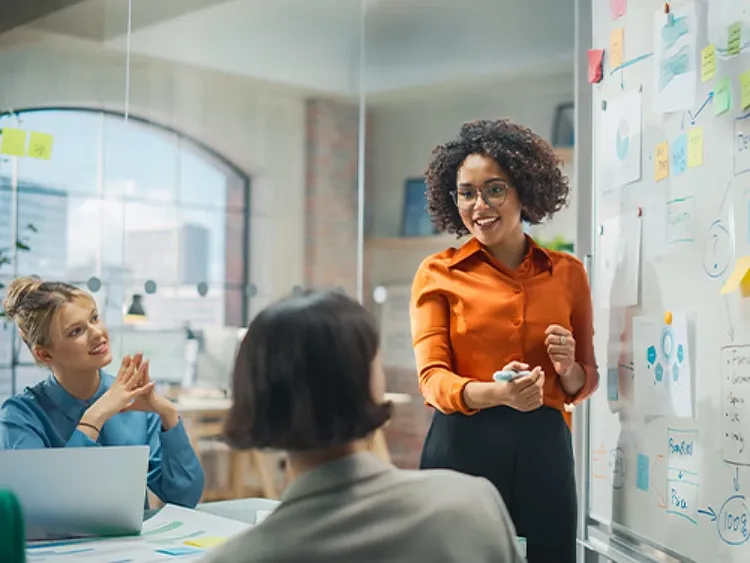 A woman standing next to a whiteboard giving presentation in front of two female colleagues