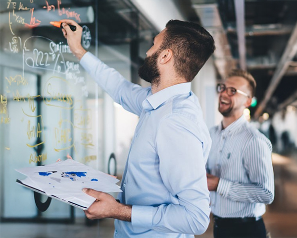 Two men collaboratively writing ideas on a transparent glass wall in a modern office setting.