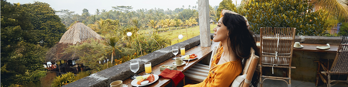 A woman on a balcony overlooks a verdant landscape, surrounded by trees and rolling hills under a clear sky.
