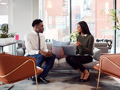 Young woman sitting on a couch with an open laptop & talking to a young professional man