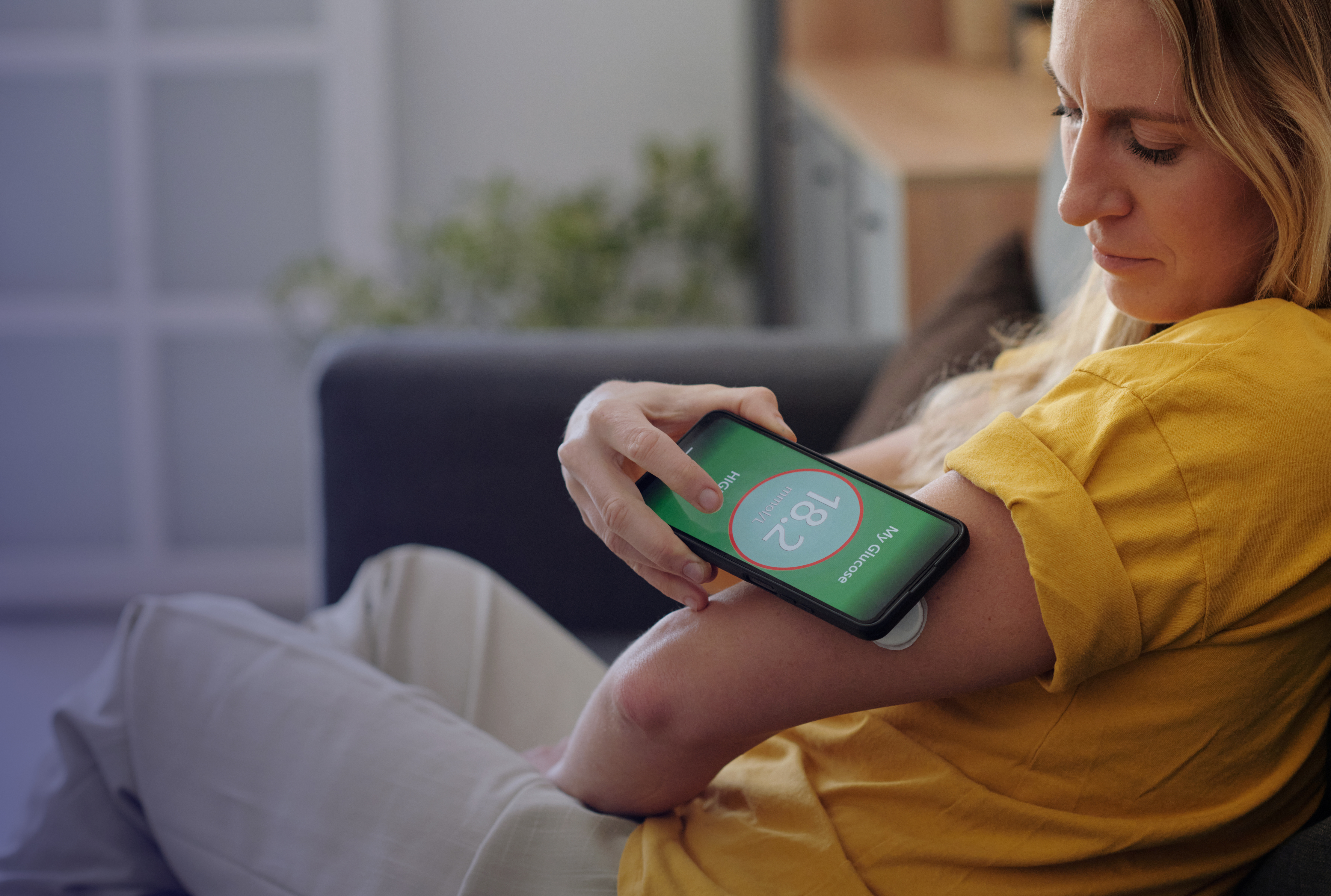 Side view closeup of woman checking glucose levels on phone with monitoring device on arm while sitting on cozy couch copy space