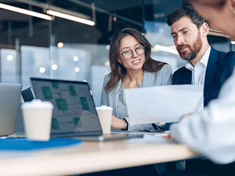 Two colleagues sitting on a meeting room chairs and looking into a laptop and having a discussion while a third colleague is sitting on the side writing.