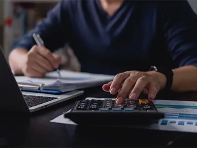 A person sitting in front of a laptop while working on a calculator next to it and making notes