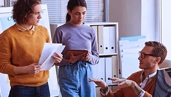 A couple of young employees standing and listening to a senior person sitting and talking to them