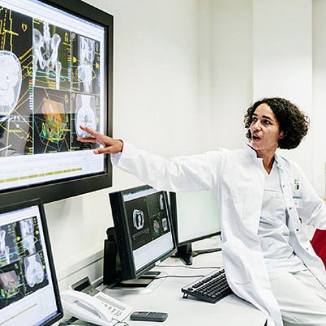 A medical professional, perched on a workdesk, pointing at a screen on the wall and talking