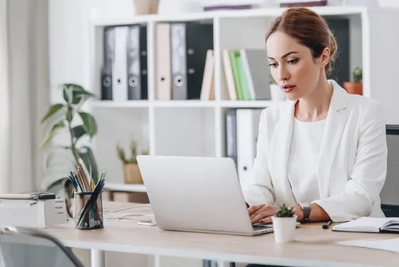 A woman in a white suit is focused on her laptop, working intently in a modern office setting.