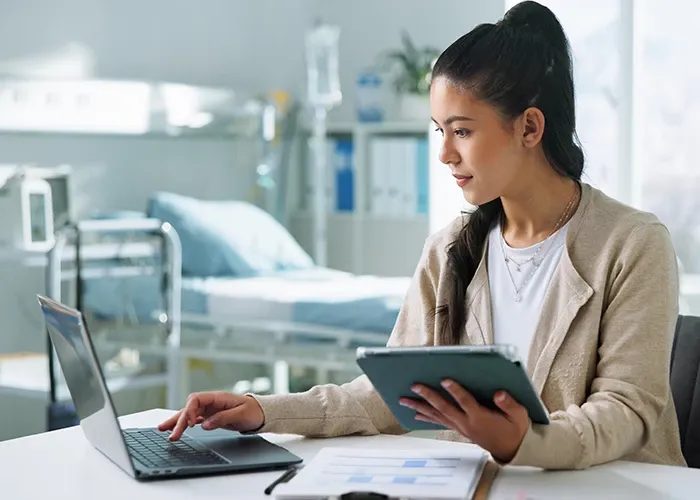A woman holding a tablet in one hand while typing on laptop with the other while sitting on a table.