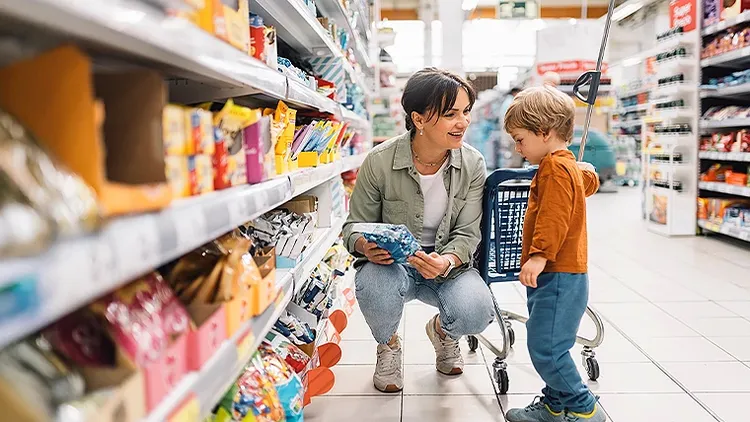 A lady sitting on her haunches in a retail store aisle, holding a package and talking to a child.