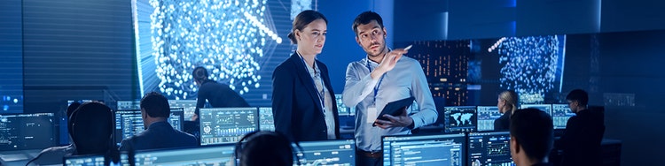 A man holding a tablet is pointing at something with his finger while a female colleague watches in a control room with many monitors