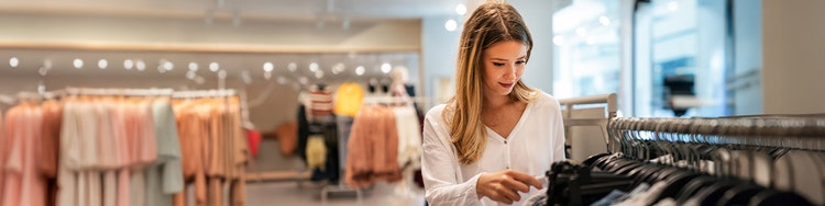 Person browsing clothing racks inside a brightly lit retail store, selecting garments from a display of neatly arranged apparel, with additional clothing sections visible in the background.