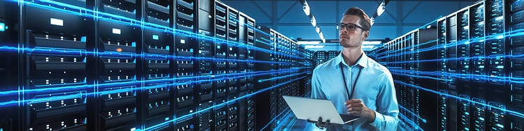 A man stands in a server room, surrounded by rows of servers and blinking lights, focused on his work.
