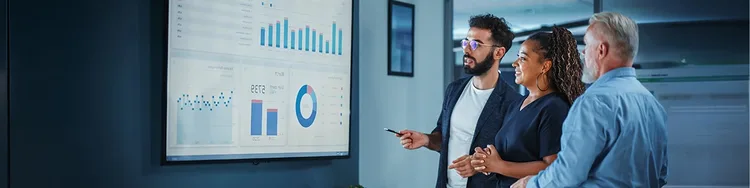 Three business professionals analyze graphs displayed on a large screen during a meeting.
