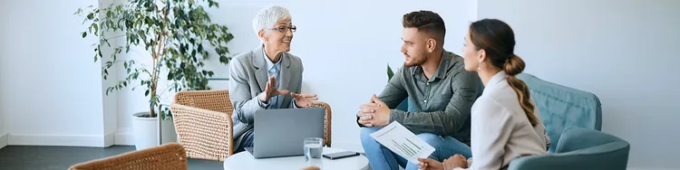 Three individuals seated in a meeting room, engaged in discussion with a laptop open on the table.
