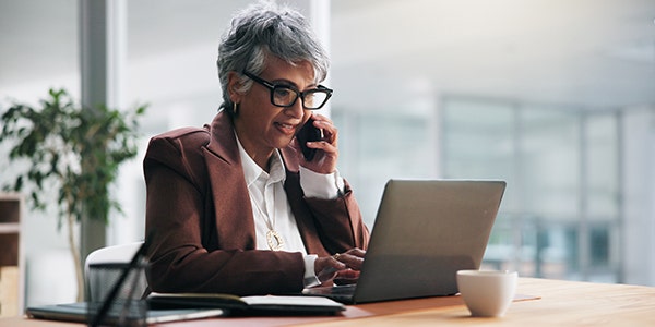 A professional woman talking on a phone while typing on a laptop with the other hand