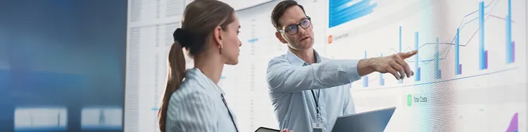 A man and woman analyze graphs displayed on a screen, engaged in a discussion about the data presented.