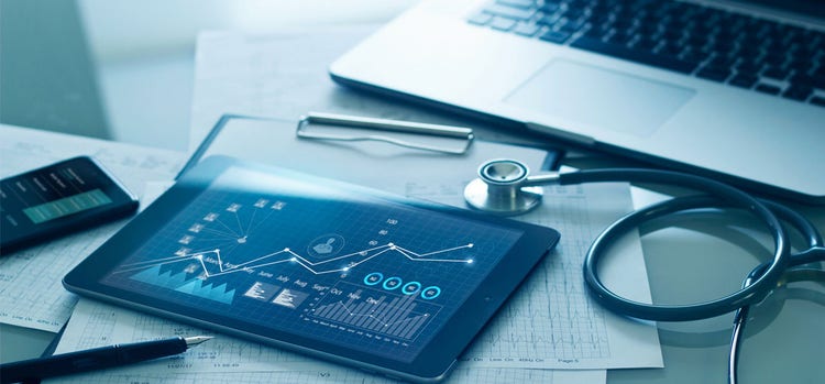 A tab, pen, mobile, laptop, stethoscope and scan reports are placed on a glass table.