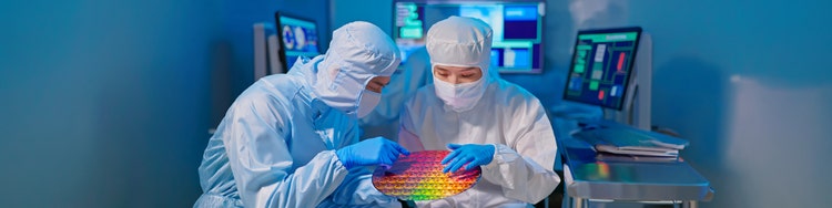 Two scientists in cleanroom suits examine a colorful silicon wafer in a high-tech lab, with monitors displaying data in the background.
