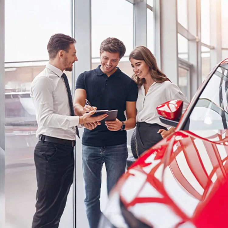 A male and female couple and a salesman looking at a tablet while standing by a new car.