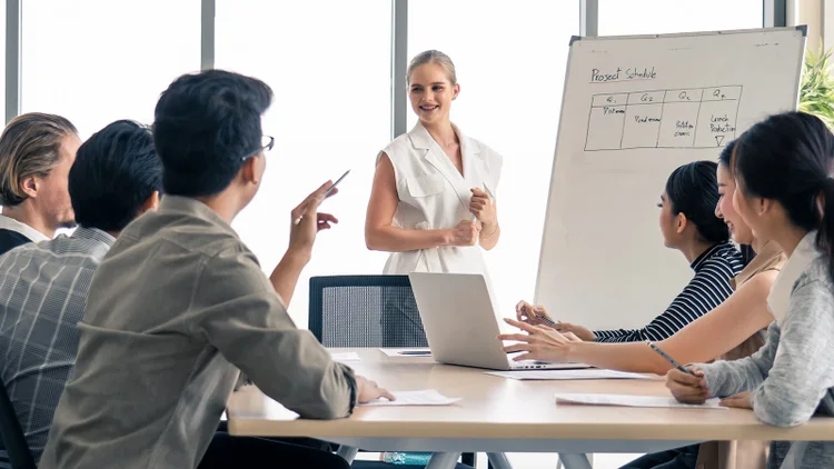A woman presents a project schedule on a whiteboard to colleagues sitting at a table