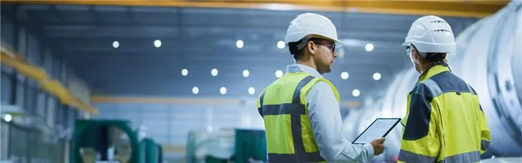 Two male colleagues in hard hat and safety vest having a discussion while looking at a tablet.