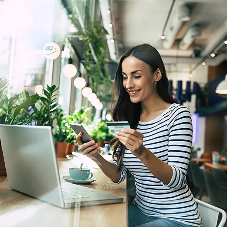 A woman sitting in front of her laptop with a smartphone in one hand and a card in the other
