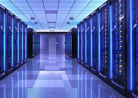 A data center corridor lit in blue, lined with tall server racks on both sides leading to a door at the far end.